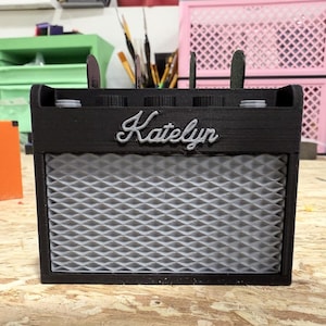 May include: A black, miniature amplifier with the name "Katelyn" in cursive script across the front. The speaker grill is a light gray, diamond-patterned design. The top has several knobs and slots for holding items.