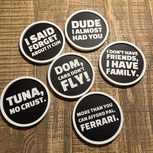 May include: A collection of six round black coasters with white rims and humorous white text. The phrases include "I SAID FORGET ABOUT IT CUH," "DUDE I ALMOST HAD YOU," and "DOM, CARS DON'T FLY!" Other coasters say "TUNA, NO CRUST," "I DON'T HAVE FRIENDS, I HAVE FAMILY," and "MORE THAN YOU CAN AFFORD PAL, FERRARI."