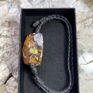 May include: A bracelet featuring a polished, multi-colored boulder opal gemstone with brown, tan, and iridescent green and orange flecks. The stone is secured by a black cord with a braided design and adjustable knots, presented in a black box.