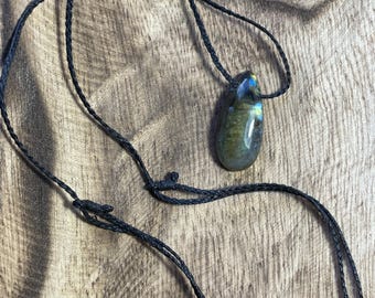 May include: A teardrop-shaped labradorite pendant featuring iridescent blue and gold flashes, suspended from a thin, braided black cord necklace. The polished stone displays a smooth, reflective surface with natural color variations throughout the gem.