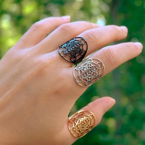 May include: Three rings featuring a geometric Metatron's Cube design. One ring is matte black, one is polished silver-tone, and one is polished gold-tone. Each ring has a wide, circular band with an intricate, openwork pattern of intersecting lines.