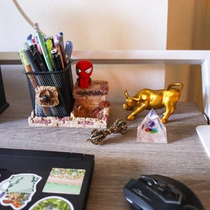 May include: A desk workspace with a black laptop, a black computer mouse, a gold bull figurine, a clear crystal pyramid, a gold metal decorative orb, a red and black cartoon figurine, a black wire mesh pencil holder with pencils and pens, and a sticker with the word "ELEMENT" on a white background.