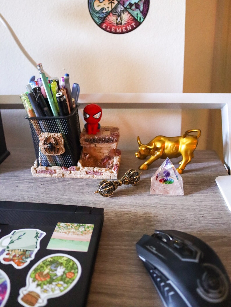 May include: A desk workspace with a black laptop, a black computer mouse, a gold bull figurine, a clear crystal pyramid, a gold metal decorative orb, a red and black cartoon figurine, a black wire mesh pencil holder with pencils and pens, and a sticker with the word "ELEMENT" on a white background.