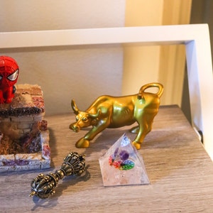 May include: A gold bull figurine, a clear crystal pyramid with a rainbow and purple stone inside, and a gold-colored metal hand-held prayer wheel on a wooden surface.
