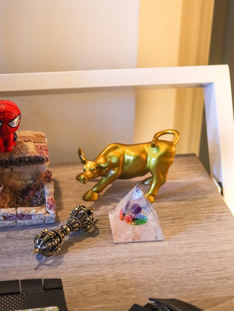 May include: A gold bull figurine, a clear crystal pyramid with a rainbow and purple stone inside, and a gold-colored metal hand-held prayer wheel on a wooden surface.