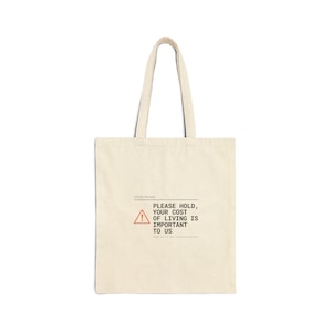 May include: Cream-colored tote bag with a rectangular design. The bag features a printed message in black font with a red warning symbol. The text reads: "PLEASE HOLD, YOUR COST OF LIVING IS IMPORTANT TO US."