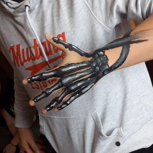 May include: A person's arm with a painted skeleton hand. The hand is painted in black and grey, with the bones visible. The person is wearing a grey sweatshirt with the word "Mustang" printed on it.