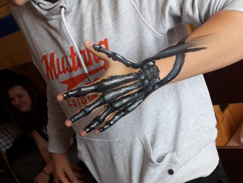 May include: A person's arm with a painted skeleton hand. The hand is painted in black and gray, with the bones visible. The person is wearing a gray sweatshirt with the word "Mustang" printed on it.