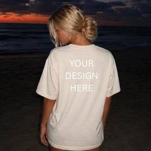 May include: Cream-colored t-shirt with the text "YOUR DESIGN HERE" in white block letters. The shirt is worn by a person on a beach at dusk, with the ocean and sunset in the background.