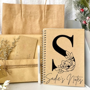 May include: A brown kraft paper notebook with a black floral design and the letter "S" on the front. The notebook has a spiral binding and the text "Sadie's Notes" is printed on the front.