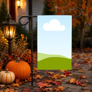 Könnte beinhalten: Eine rechteckige Gartenflagge mit einem einfachen grafischen Design aus einer einzelnen weißen Wolke vor hellblauem Himmel über grünen Hügeln, aufgehängt an einem schwarzen Metallständer mit gebogenen Enden.
