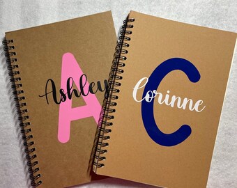 May include: Two brown spiral notebooks with personalized names. The notebook on the left has a pink "A" for "Ashley" and the notebook on the right has a blue "C" for "Corinne".