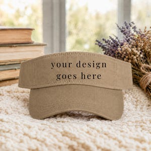 May include: A tan-colored cotton visor featuring the black text "your design goes here" printed on the front panel. The visor has a curved brim and a structured crown, designed to be worn as headwear for sun protection and outdoor activities.