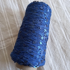 Op de afbeelding: Een spoel donkerblauw garen met ingebedde pailletten. Het garen is gewikkeld rond een kartonnen koker. De pailletten, in een donkerdere blauwtint, zijn verspreid over het garen. De spoel ligt op een licht, gestreept oppervlak.
