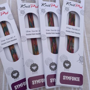 Könnte beinhalten: Vier Packungen Knit Pro Symfonie Doppelspitzen-Stricknadeln. Die mehrfarbigen Nadeln sind durch ein ausgestanztes Fenster sichtbar. Jede Packung zeigt die Nadelgröße in Millimetern und Zoll an. Die Verpackungen zeigen ein Schaf-Logo und den Markennamen.