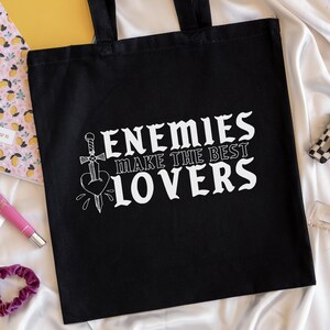 May include: Black tote bag with white text that reads "ENEMIES MAKE THE BEST LOVERS". The design includes a sword piercing a heart. The bag has two black handles and is made of a sturdy fabric.