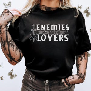 May include: Black t-shirt with the text "ENEMIES MAKE THE BEST LOVERS" in white. The design includes a sword piercing a heart. The shirt is a crew neck style.