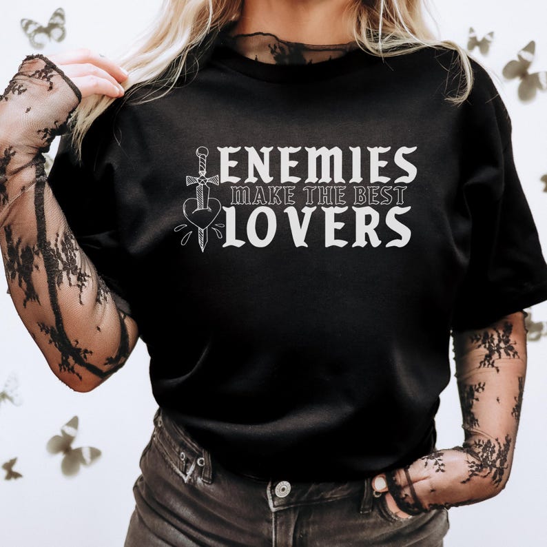May include: Black t-shirt with the text "ENEMIES MAKE THE BEST LOVERS" in white. The design includes a sword piercing a heart. The shirt is a crew neck style.