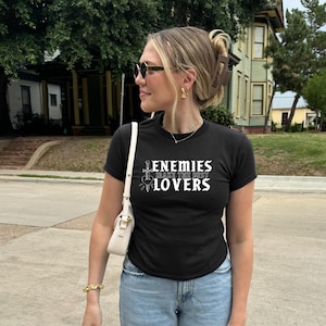 May include: Black crew neck t-shirt with the text "ENEMIES MAKE THE BEST LOVERS" in white. The shirt features a sword and heart graphic. The person is wearing sunglasses, gold earrings, and a white purse.
