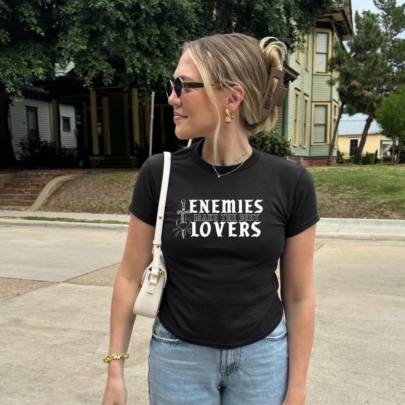 May include: Black crew neck t-shirt with the text "ENEMIES MAKE THE BEST LOVERS" in white. The shirt features a sword and heart graphic. The person is wearing sunglasses, gold earrings, and a white purse.