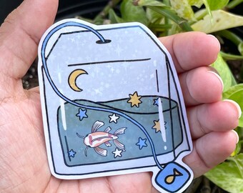 May include: A blue tea bag with a white string and a blue tag. Inside the tea bag is a clear container with a blue liquid, a white and orange fish, and yellow and white stars.
