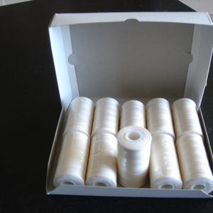 May include: A white cardboard box containing several spools of off-white thread. The spools are arranged inside the box, with the thread wound tightly around each spool. The box is on a dark surface.
