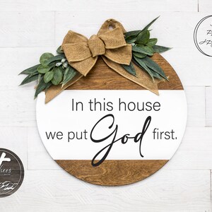 May include: A round wooden sign with a white background and a brown wood grain border. The sign has a burlap bow and greenery on top. The text on the sign reads "In this house we put God first."