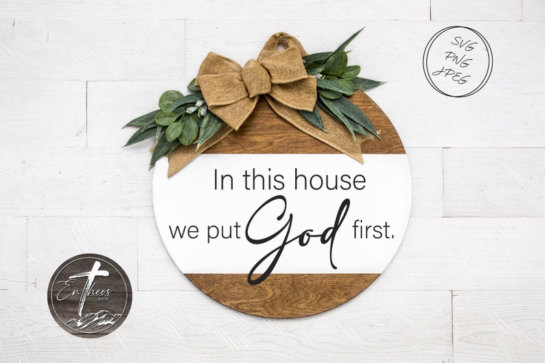 May include: A round wooden sign with a white background and a brown wood grain border. The sign has a burlap bow and greenery on top. The text on the sign reads "In this house we put God first."