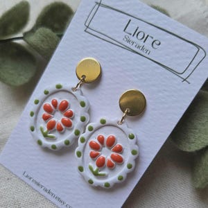 Op de afbeelding: Een paar witte bloemen oorbellen met oranje bloemblaadjes en groene bladeren. De oorbellen hebben een geschulpte rand met groene stippen en goudkleurige ronde studs. De oorbellen worden gepresenteerd op een witte kaart met de tekst "Liore Sieraden".