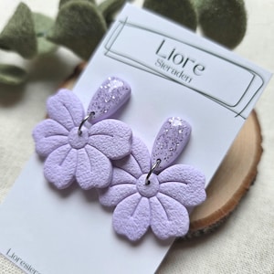 Op de afbeelding: Een paar lavendelkleurige bloemvormige oorbellen met een getextureerde afwerking. De oorbellen hebben een zilveren glitteraccent op het bovenste gedeelte en zijn bevestigd aan een witte kaart met de tekst "Liore Steraden".