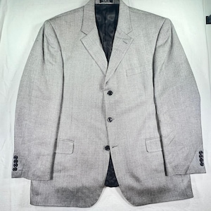 May include: A light gray blazer featuring a subtle herringbone pattern, a notched lapel, and a three-button front closure. The garment includes a chest pocket, two flap pockets at the waist, and four buttons on each sleeve cuff, with a black lining.