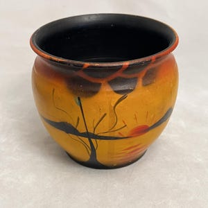 May include: A small, rounded ceramic pot featuring a hand-painted sunset scene with a black silhouette of a tree against a vibrant yellow and orange background. The interior of the pot is solid black, and the rim is accented with a thin orange line.