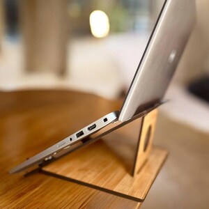 May include: A silver laptop computer is propped up on a wooden laptop stand. The laptop is angled upwards and the stand is made of light brown wood.