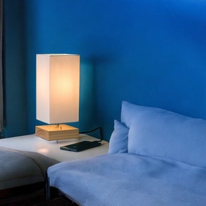 May include: A white lamp with a wooden base sits on a white bedside table next to a blue bed with a white pillow. A black phone is on the bedside table.