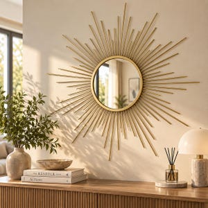 May include: A circular wall mirror with a gold-toned sunburst frame featuring thin, radiating metal spokes of varying lengths. The mirror has a smooth, reflective centre and is mounted on a plain wall, creating a star-like geometric pattern.