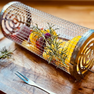 May include: A metal grilling basket containing yellow peppers, red onion, and rosemary sprigs. The basket is on a wooden chopping board with a silver fork.