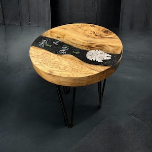May include: A round side table featuring a natural wood grain top with a central black epoxy resin river inlay. The resin section depicts a space battle scene with a Millennium Falcon, TIE fighters, and green laser beams. It has black metal legs.
