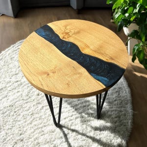 May include: Round wooden coffee table with a dark blue resin river flowing through the centre. The table has black hairpin legs.