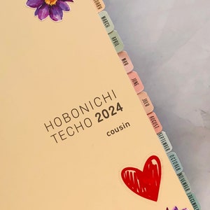 May include: A Hobonichi Techo 2024 Cousin planner with colorful monthly tabs and stickers. The planner has a light beige cover with the text "HOBONICHI TECHO 2024" and "cousin" printed on it.