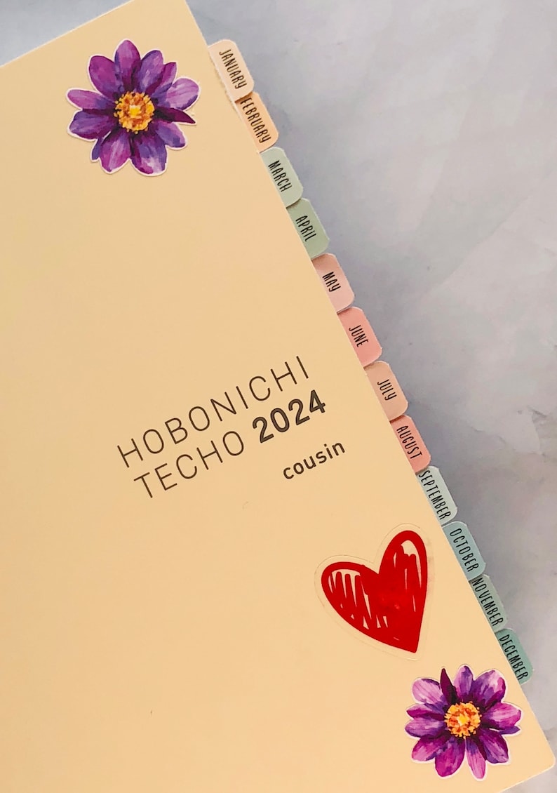May include: A Hobonichi Techo 2024 Cousin planner with colorful monthly tabs and stickers. The planner has a light beige cover with the text "HOBONICHI TECHO 2024" and "cousin" printed on it.
