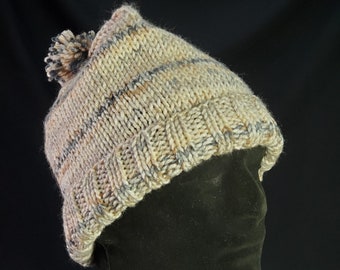 Può includere: Un berretto lavorato a mano a maglia in tonalità neutre, con bordo a coste e motivo a righe nei toni del beige, grigio e marrone. Il cappello è sormontato da un pompon abbinato. Il cappello è esposto su una testa di manichino nera.