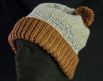 Può includere: Un berretto lavorato a maglia fatto a mano con bordo a coste marrone e corona maculata grigio chiaro e bianca. Il cappello presenta un pompon marrone abbinato sulla parte superiore. Il cappello è esposto su una testa di manichino nera su sfondo nero.