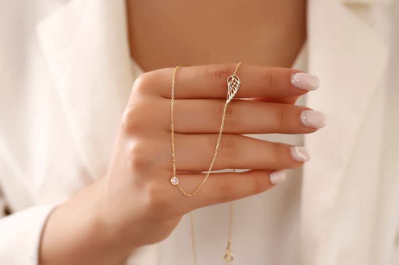 May include: A delicate gold necklace featuring a small, round, clear gemstone and a wing-shaped charm. The necklace is held up against a white background, showcasing its fine details and elegant design. The necklace is a piece of jewelry.