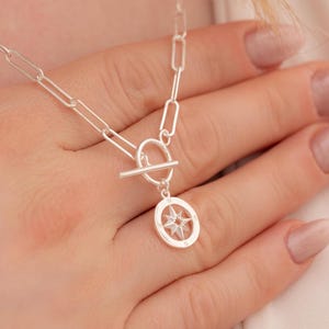 May include: A silver-colored necklace with a compass pendant. The necklace features a toggle clasp and a chain with elongated links. The compass pendant has a star design inside a circular frame. The necklace is displayed on a person's hand.