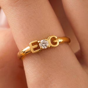 May include: A gold ring featuring the letters "E" and "G" with a small, round, clear gemstone between them. The ring is on a finger, and the gold band has a smooth, polished finish.
