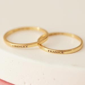 May include: Two gold-colored rings, one inscribed with "LINDSAY" and the other with "FRANKIE". The rings are interlocked, suggesting a connection. The background is a soft, neutral color, highlighting the jewelry.