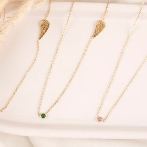 May include: Four delicate gold necklaces with different colored gemstones. Each necklace features a small, round gemstone in blue, green, and pink, and a gold wing charm. The necklaces are displayed on a white surface.