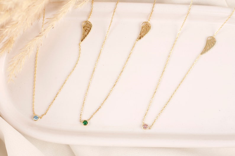 May include: Four delicate gold necklaces with different colored gemstones. Each necklace features a small, round gemstone in blue, green, and pink, and a gold wing charm. The necklaces are displayed on a white surface.