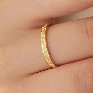 May include: A close-up of a gold ring with a repeating sunburst pattern. The ring is worn on a finger, showcasing the intricate design and the warm, yellow color of the metal. The ring is a piece of jewelry.