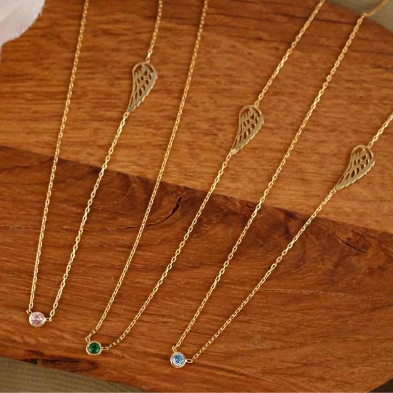May include: Four gold necklaces with delicate chains. One necklace features a pink gemstone, another a green gemstone, and a third a blue gemstone. The fourth necklace has a gold wing charm. All necklaces are displayed on a wooden surface.